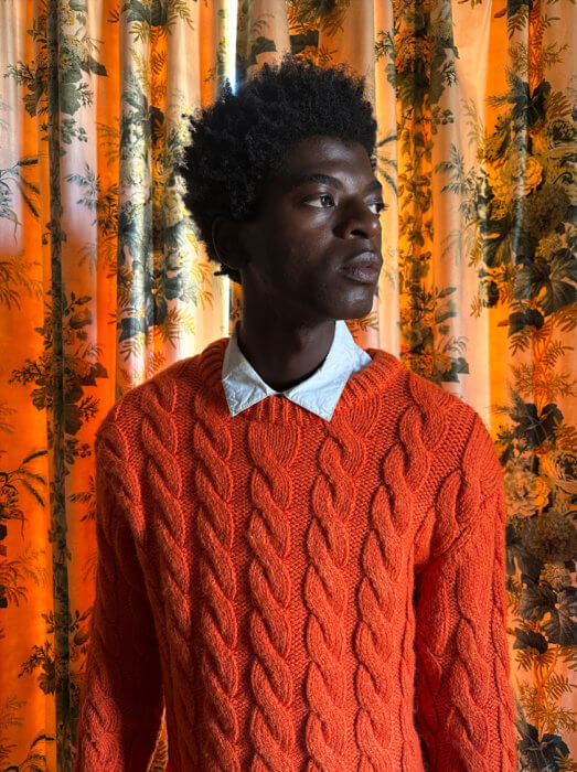 Egy mintás függöny előtt álló, élénkpiros pulóvert viselő férfit ábrázoló fotó, amely félhomályban készült a fő kamerával.