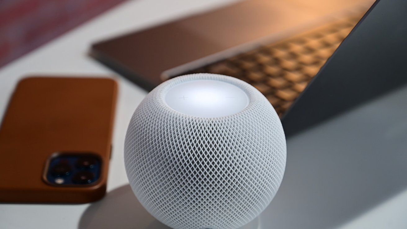 Fehér HomePod mini egy asztalon, mellette egy barna tokos iPhone-nal és egy nyitott MacBook Pro-val.
