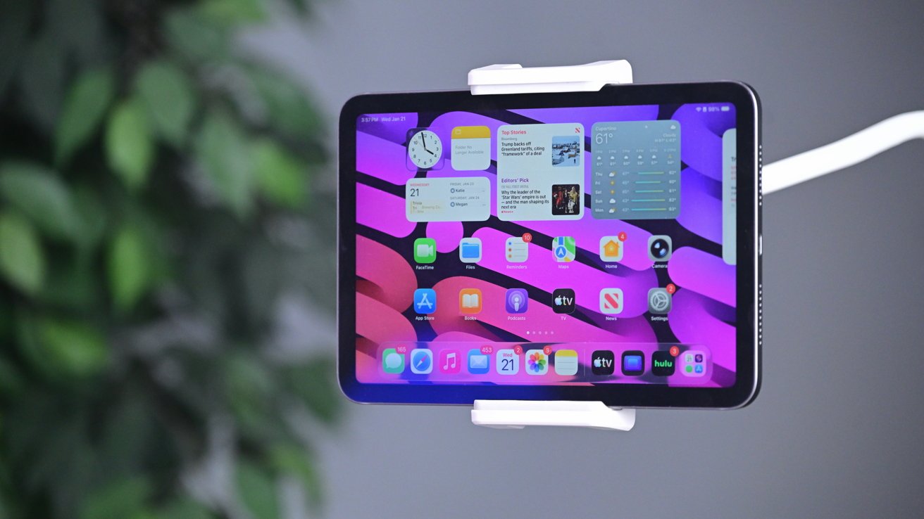 Egy iPad mini fehér csiptetős tartóban, kijelzőjén rózsaszín és lila hullámos háttérrel, alkalmazásikonokkal és widgetekkel; a háttérben elmosódott beltéri környezet és zöld növénylevelek láthatók.
